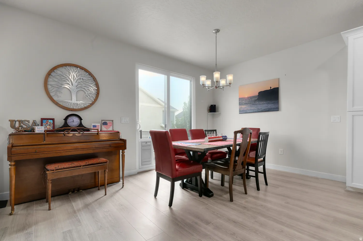 Open-concept space featuring light wood-finish flooring, a contemporary chandelier, white wall paneling, and a sliding glass door providing exterior access