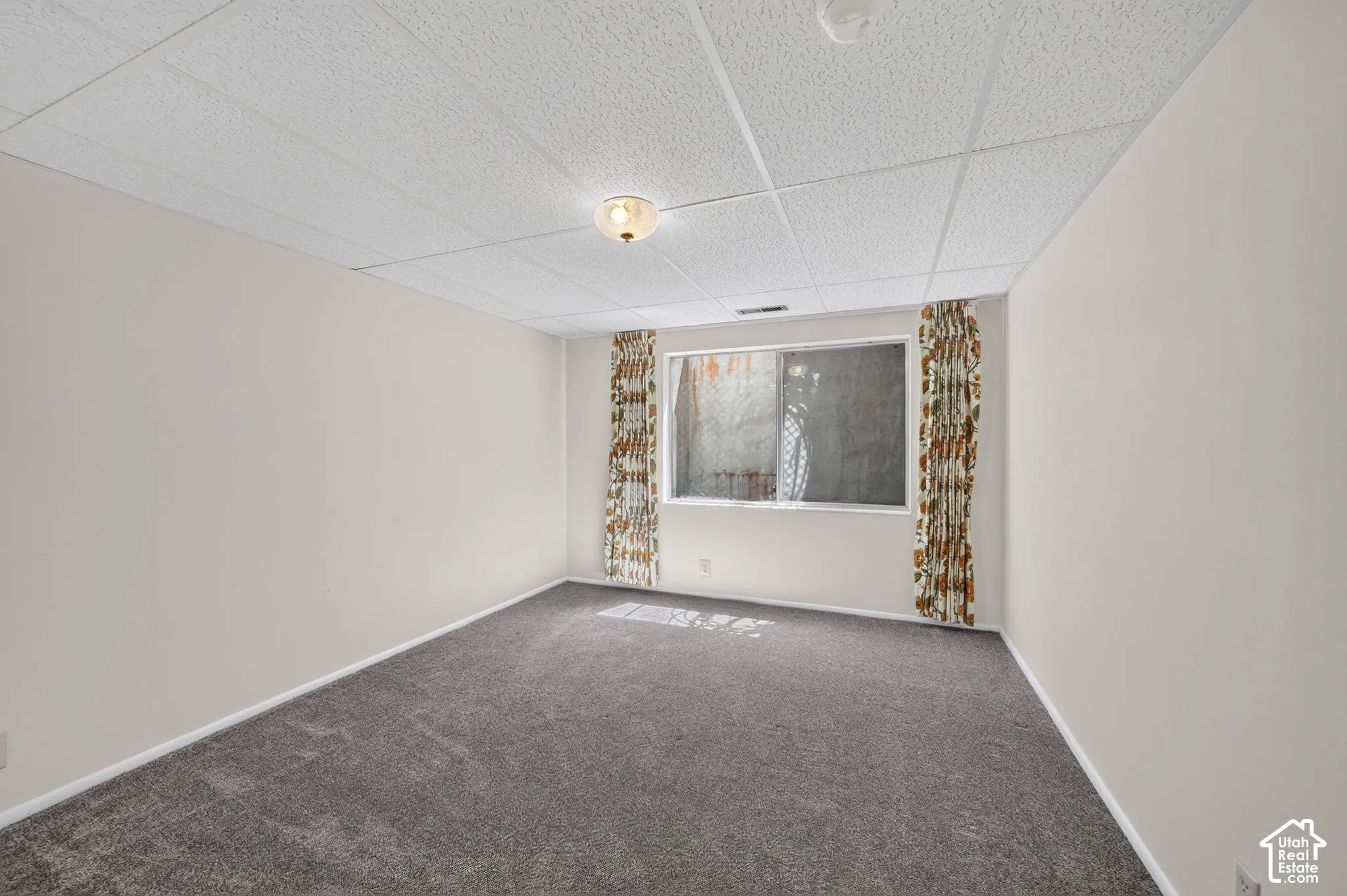 Carpeted room featuring a drop ceiling with integrated lighting, neutral wall tones, and a window with patterned drapes