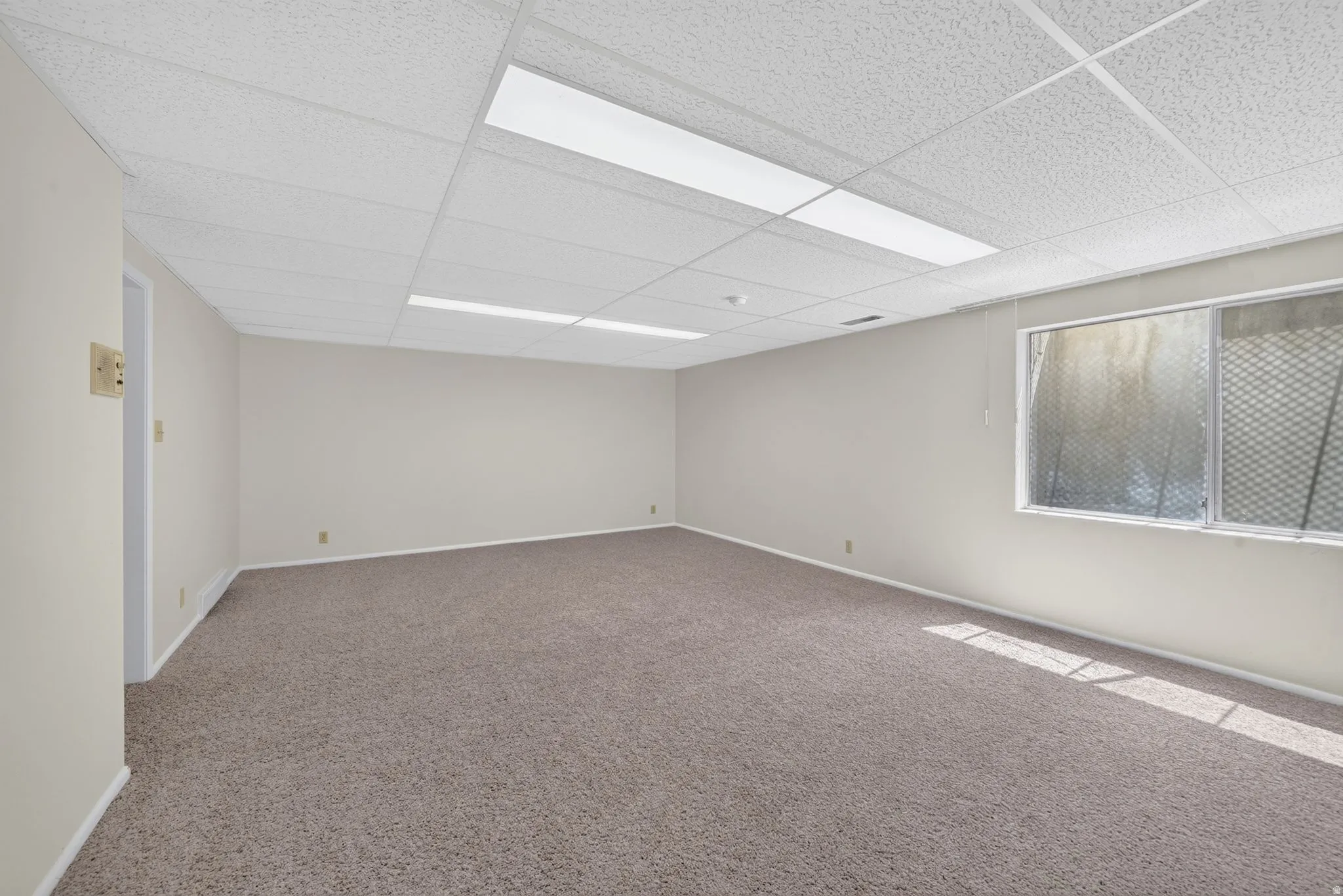 Spacious room featuring neutral carpeting, light-toned walls, and a dropped ceiling with integrated fluorescent lighting