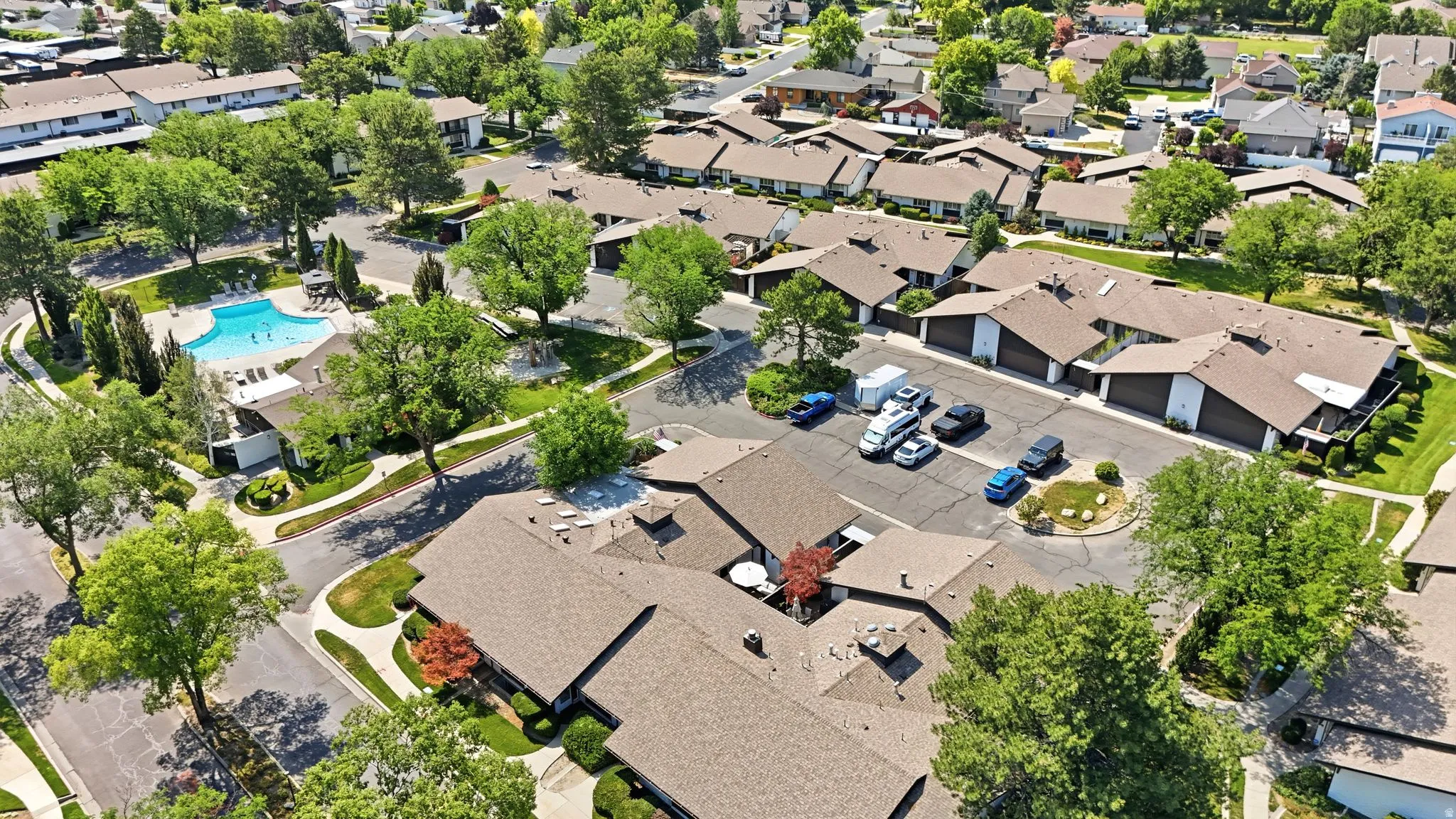 Community featuring multiple residences with brown shingle roofs, mature trees, paved roadways, and a communal swimming pool with surrounding patio area