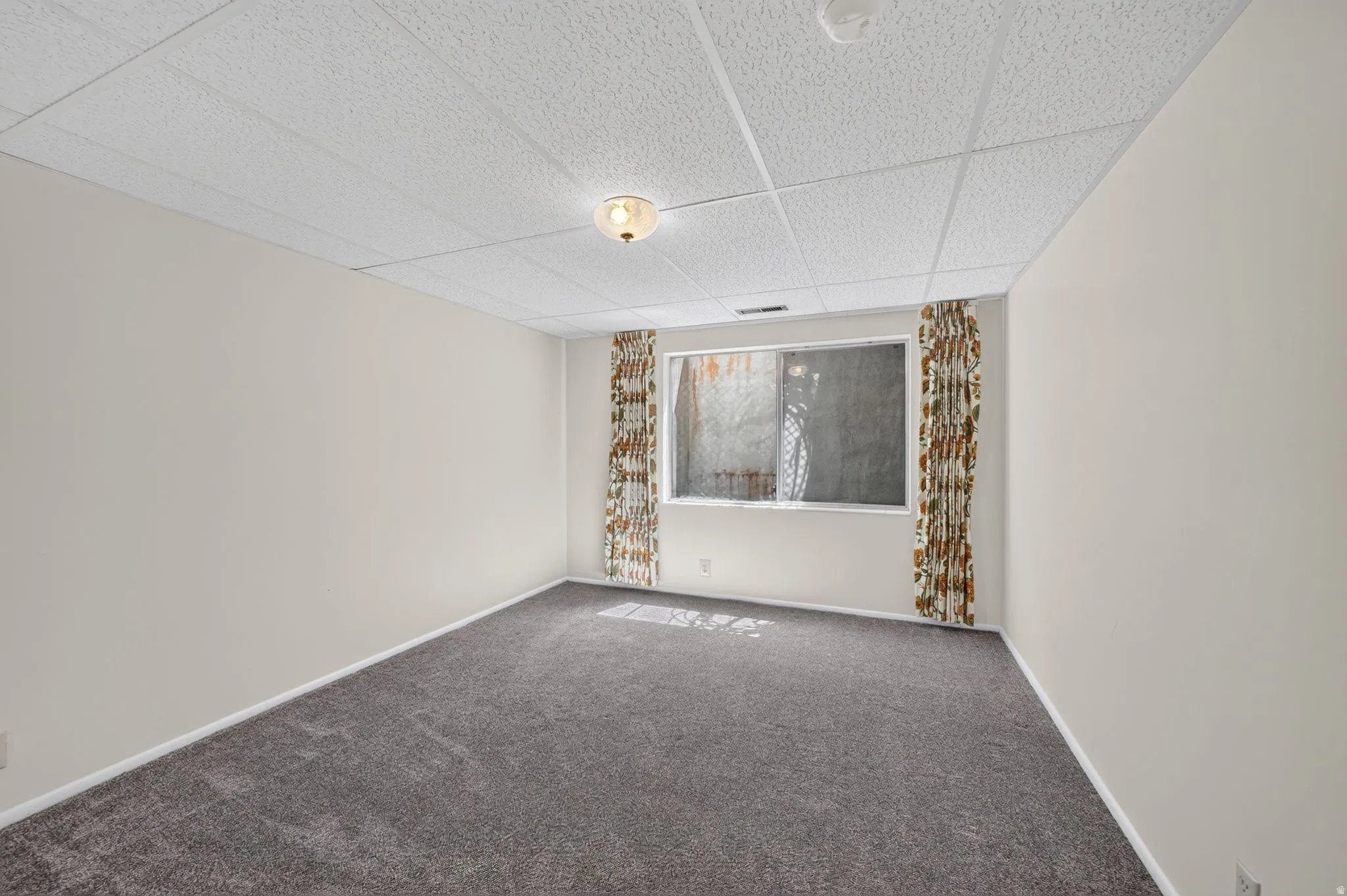 Carpeted room featuring a drop ceiling with integrated lighting, neutral wall tones, and a window with patterned drapes