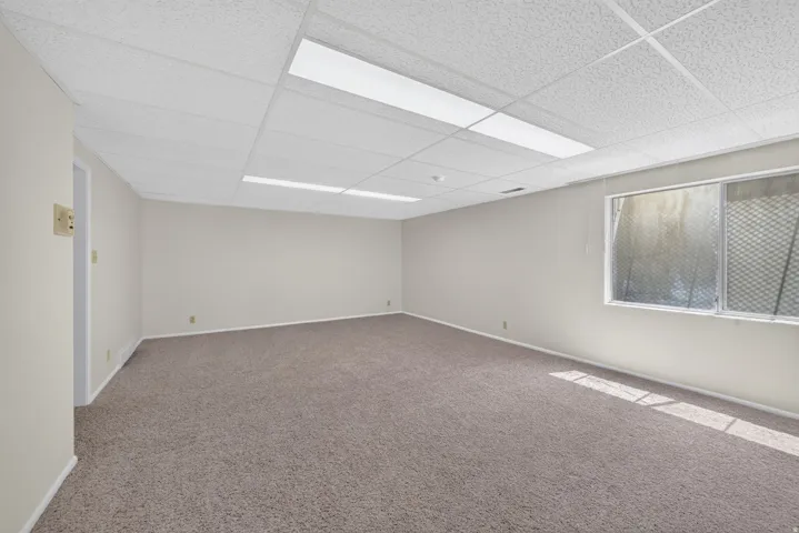 Spacious room featuring neutral carpeting, light-toned walls, and a dropped ceiling with integrated fluorescent lighting
