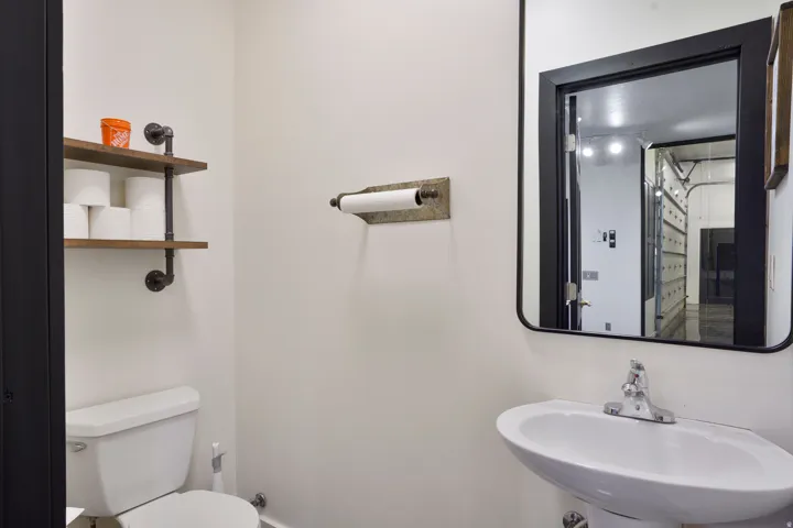 Compact powder room featuring a wall-mounted sink, a toilet, a rectangular black-framed mirror, and industrial-style wood shelves