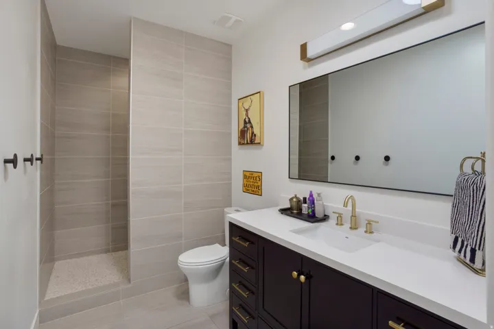 Spacious bathroom featuring a walk-in shower with light-toned wall tile, a dark wood-finish vanity with a white countertop, a rectangular undermount sink, and brass-finish fixtures