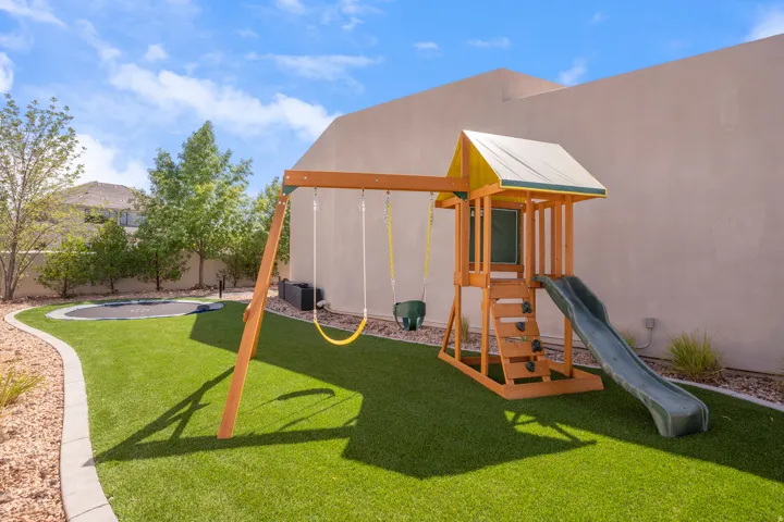 Expansive backyard featuring artificial turf, a children's playset with swings and a slide, a built-in trampoline, and mature perimeter landscaping