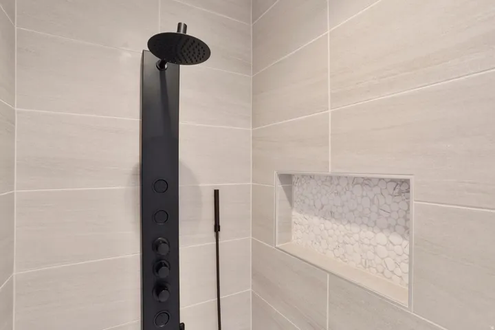 Shower enclosure featuring light-toned large format tile, a matte black multi-function shower panel with rain shower head, and an inset niche with pebble tile accent