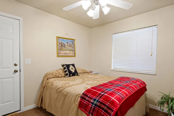 White paneled door with brass hardware, neutral wall paint, ceiling fan with light fixture, window with blinds, and carpeted flooring