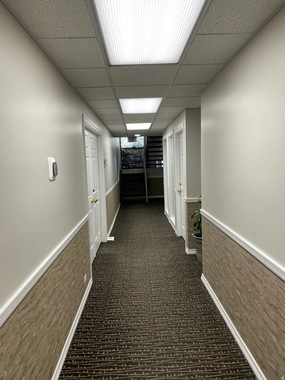 Hallway featuring patterned carpeting, wainscoting, white baseboards, and a dropped ceiling with recessed lighting