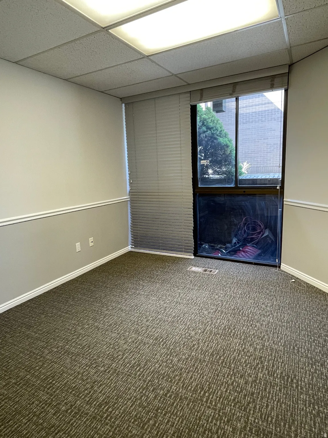 Windowed room featuring textured carpeting, white baseboards, chair rail molding, and a drop ceiling with integrated fluorescent lighting