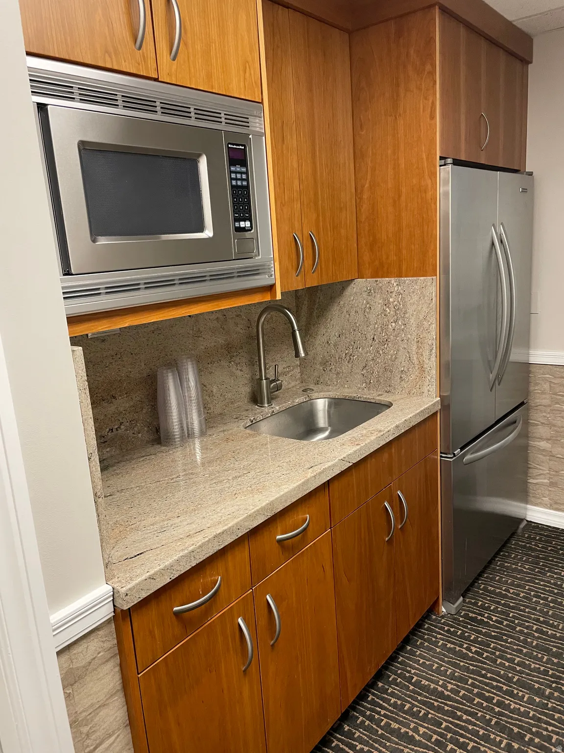 Compact kitchen area featuring wood cabinetry, a stainless steel microwave, a built-in sink with a gooseneck faucet, light-toned stone countertops, and a full-size stainless steel refrigerator