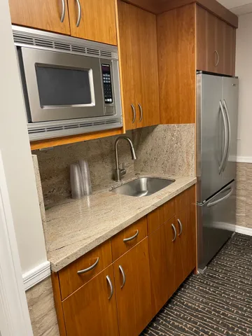Compact kitchen area featuring wood cabinetry, a stainless steel microwave, a built-in sink with a gooseneck faucet, light-toned stone countertops, and a full-size stainless steel refrigerator