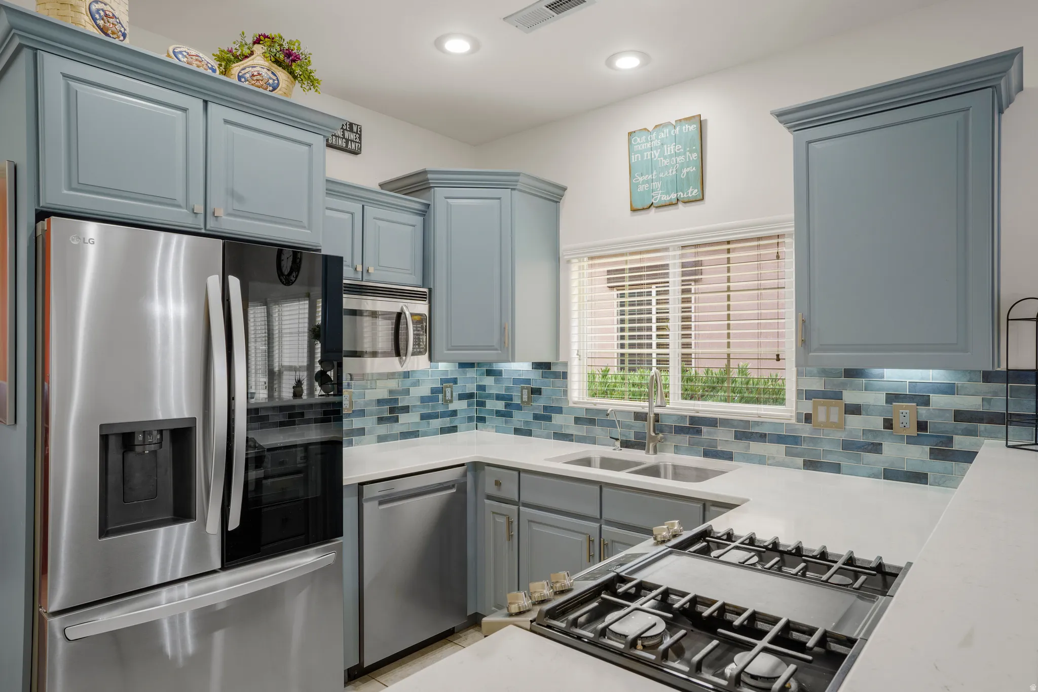 Kitchen featuring light blue cabinetry, stainless steel appliances, a tile backsplash with blue and white tones, white countertops, and a window with blinds