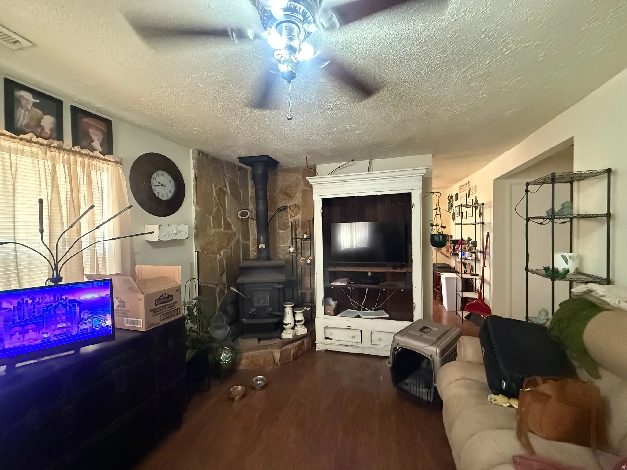 Living area featuring wood-finish flooring, a stone-clad wall with a wood-burning stove, a ceiling fan with integrated lighting, and a built-in media cabinet