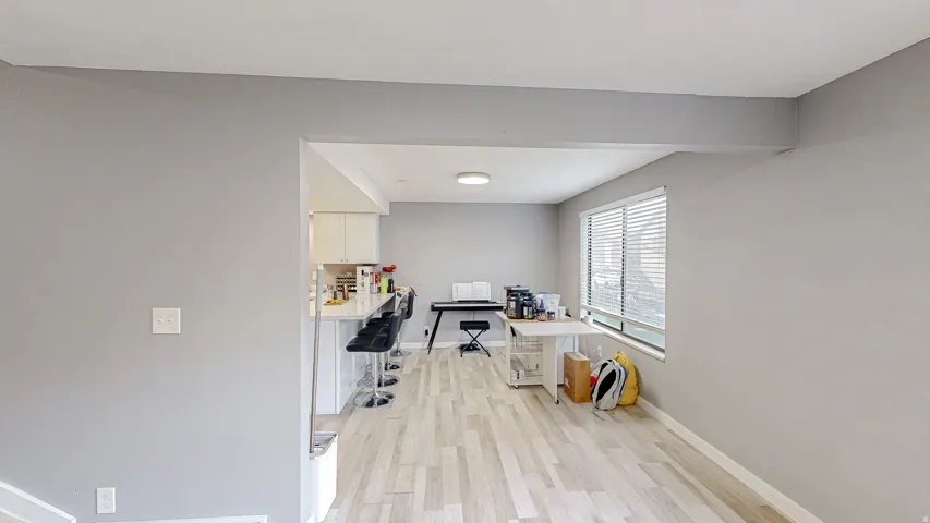 Open-concept living area featuring light wood-finish flooring, a kitchen counter bar, window with blinds, recessed ceiling lighting, and light gray wall finishes