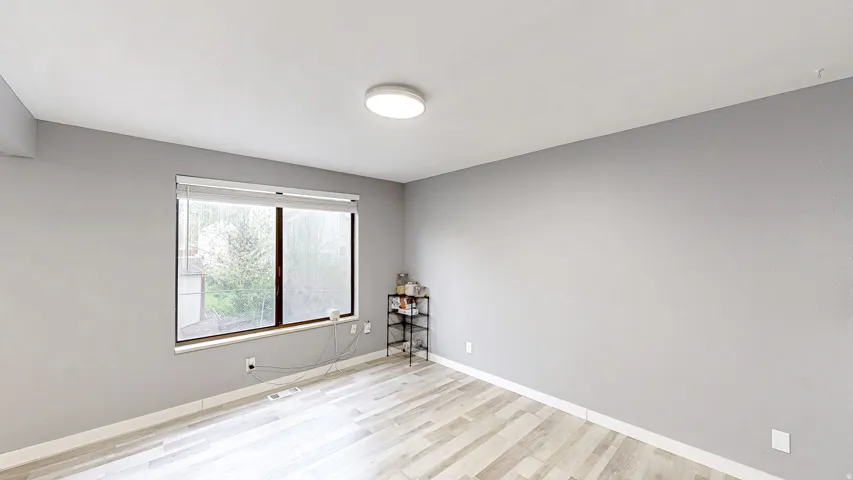 Bright interior space featuring a large window with white blinds, light wood-finish flooring, a contemporary ceiling light fixture, and neutral gray walls with white baseboards