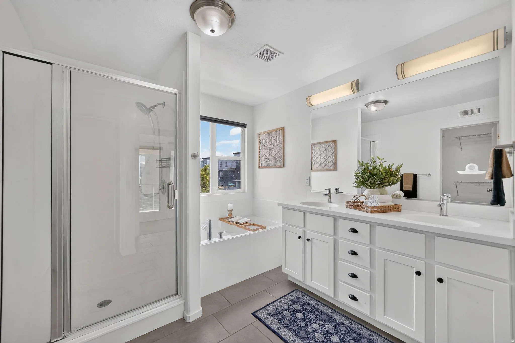 Bathroom featuring a glass-enclosed shower, a built-in bathtub with a window, a dual vanity with white cabinetry, and large format floor tiles
