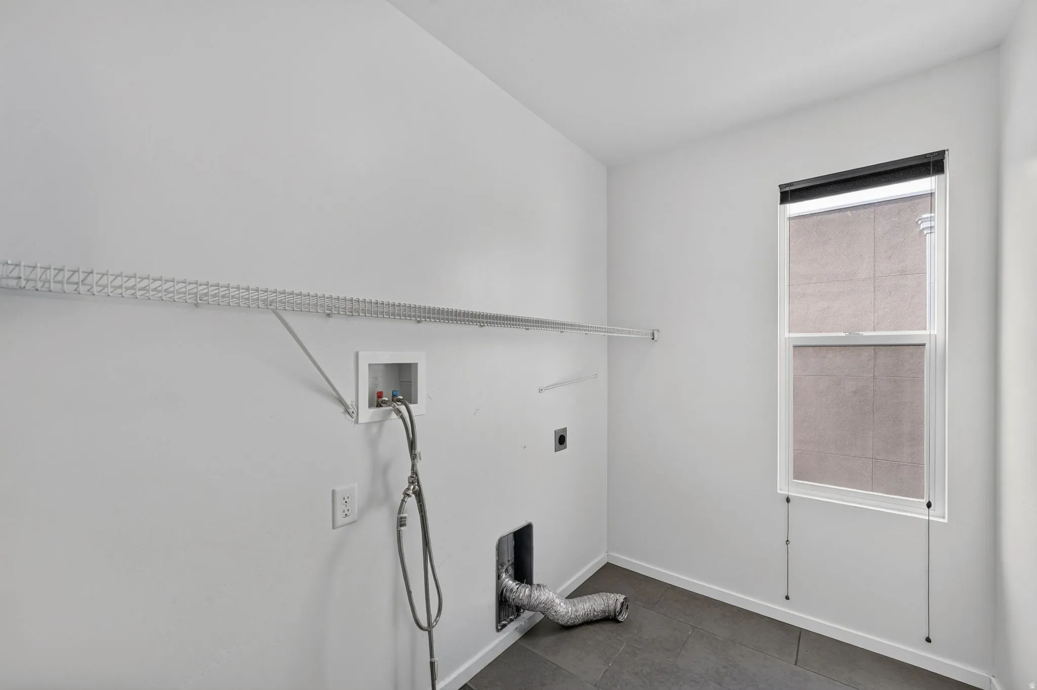 Utility room featuring a single window with roller shade, tile flooring, wire shelving, and appliance hookups