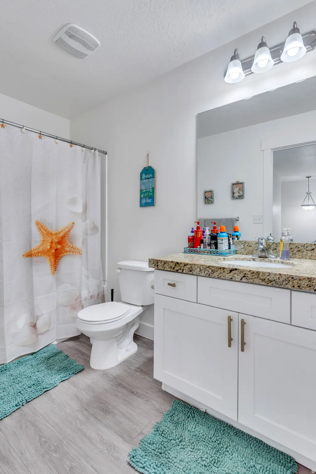 Bathroom featuring a white vanity with gold-tone hardware and a granite countertop, light wood-finish flooring, a white toilet, and a vanity mirror with a four-light fixture