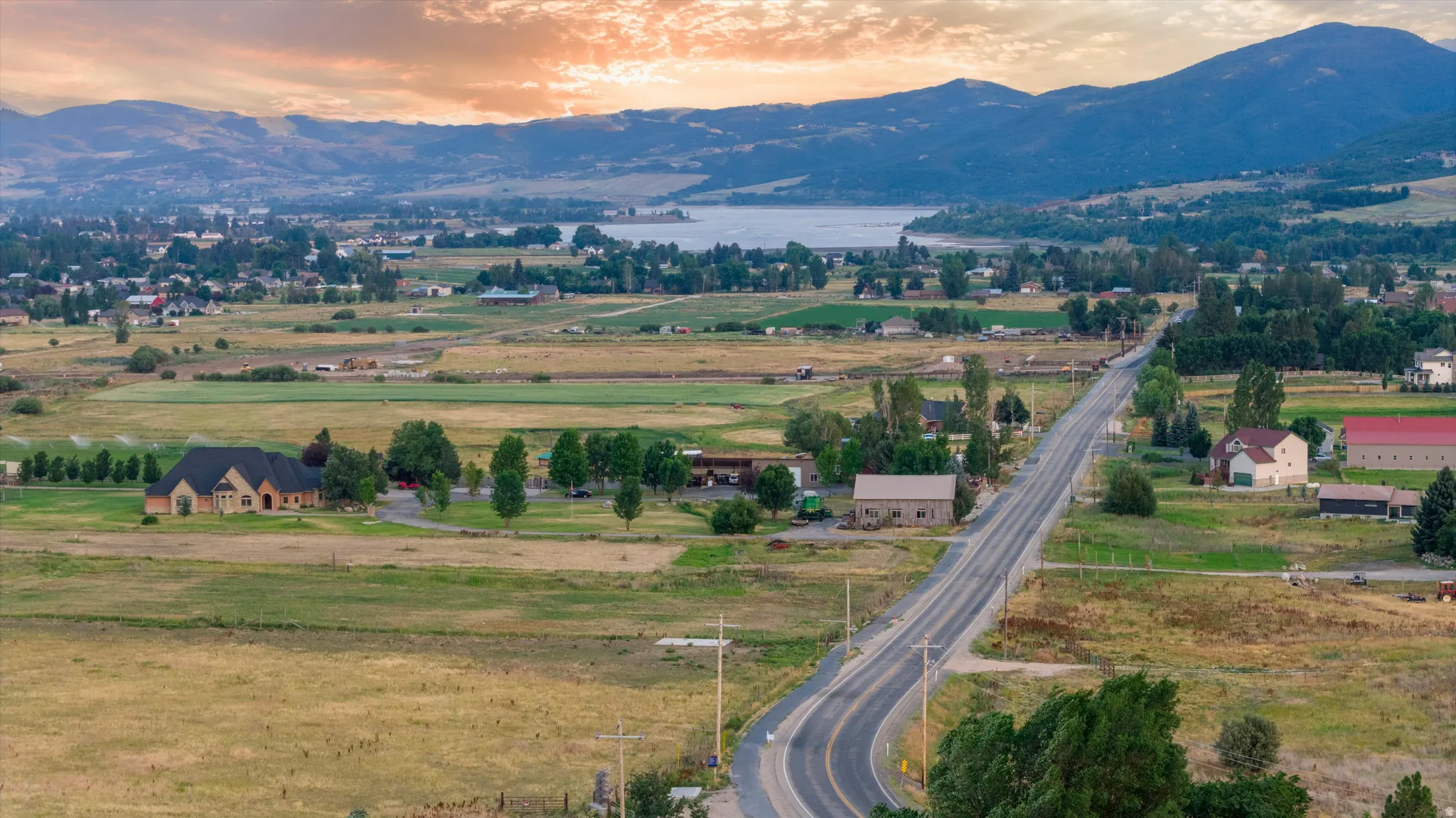 Expansive landscape featuring a paved road, open fields, distant residential structures, and a body of water with mountains in the background