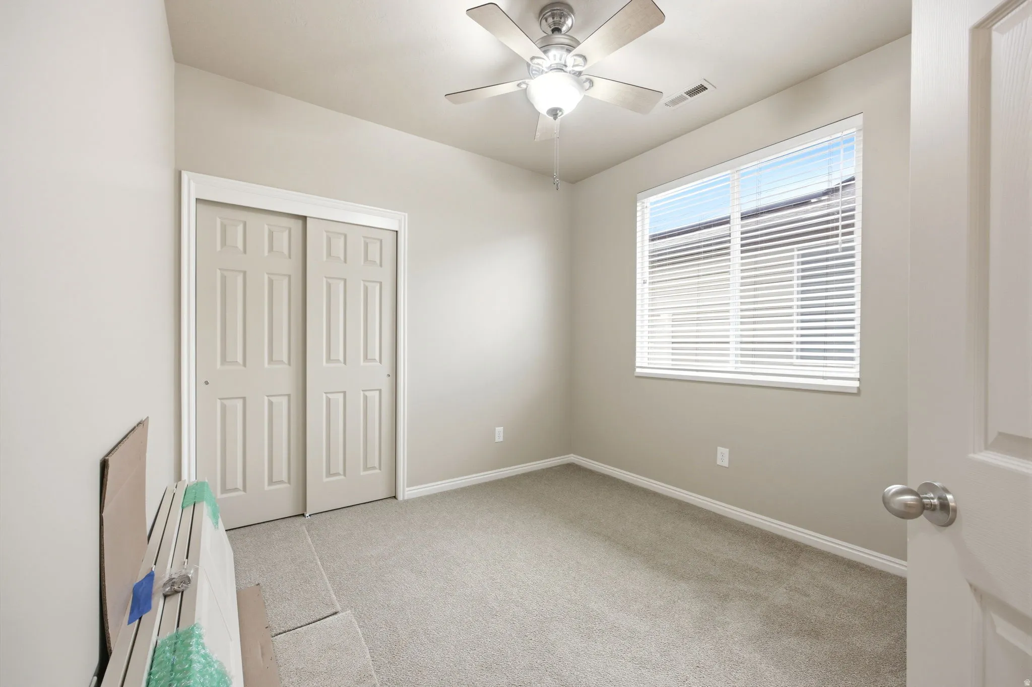 Neutral-toned room featuring a ceiling fan with light fixture, large window with blinds, and light gray wall-to-wall carpeting