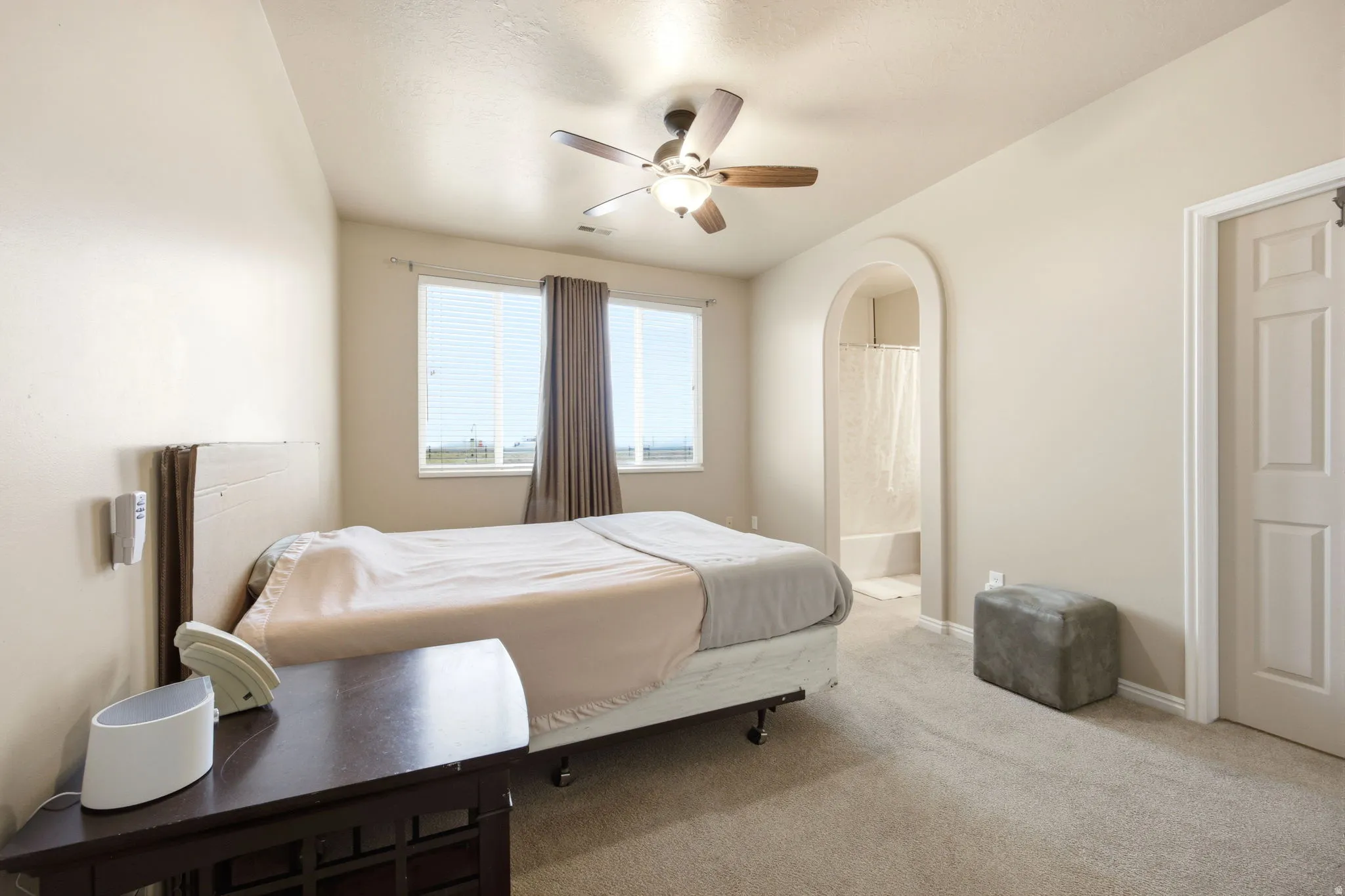 Carpeted bedroom with a ceiling fan light fixture, arched doorway to a bathroom, and a window with blinds and drapes