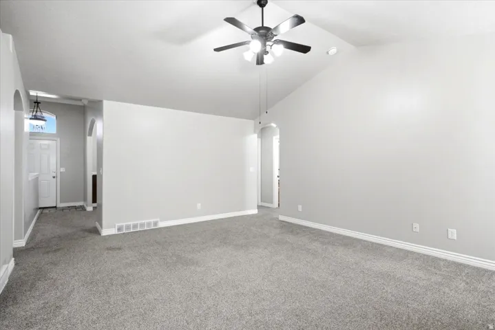 Spacious interior room featuring vaulted ceilings, a ceiling fan with integrated lighting, light gray wall paint, and gray carpet flooring