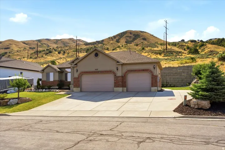 Stucco and brick exterior with a two-car garage, concrete driveway, and front lawn landscaping featuring mature trees and shrubs