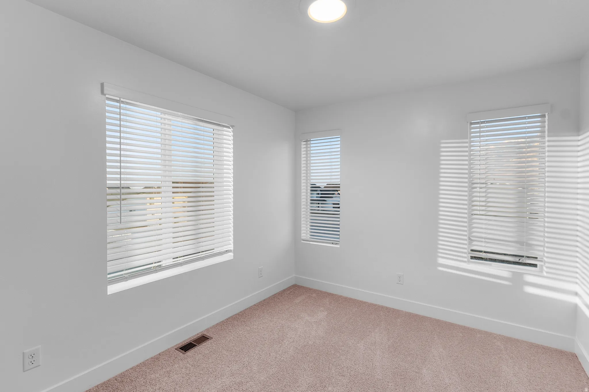 Room featuring three windows with horizontal blinds, neutral carpet flooring, white walls, and a flush mount ceiling light fixture