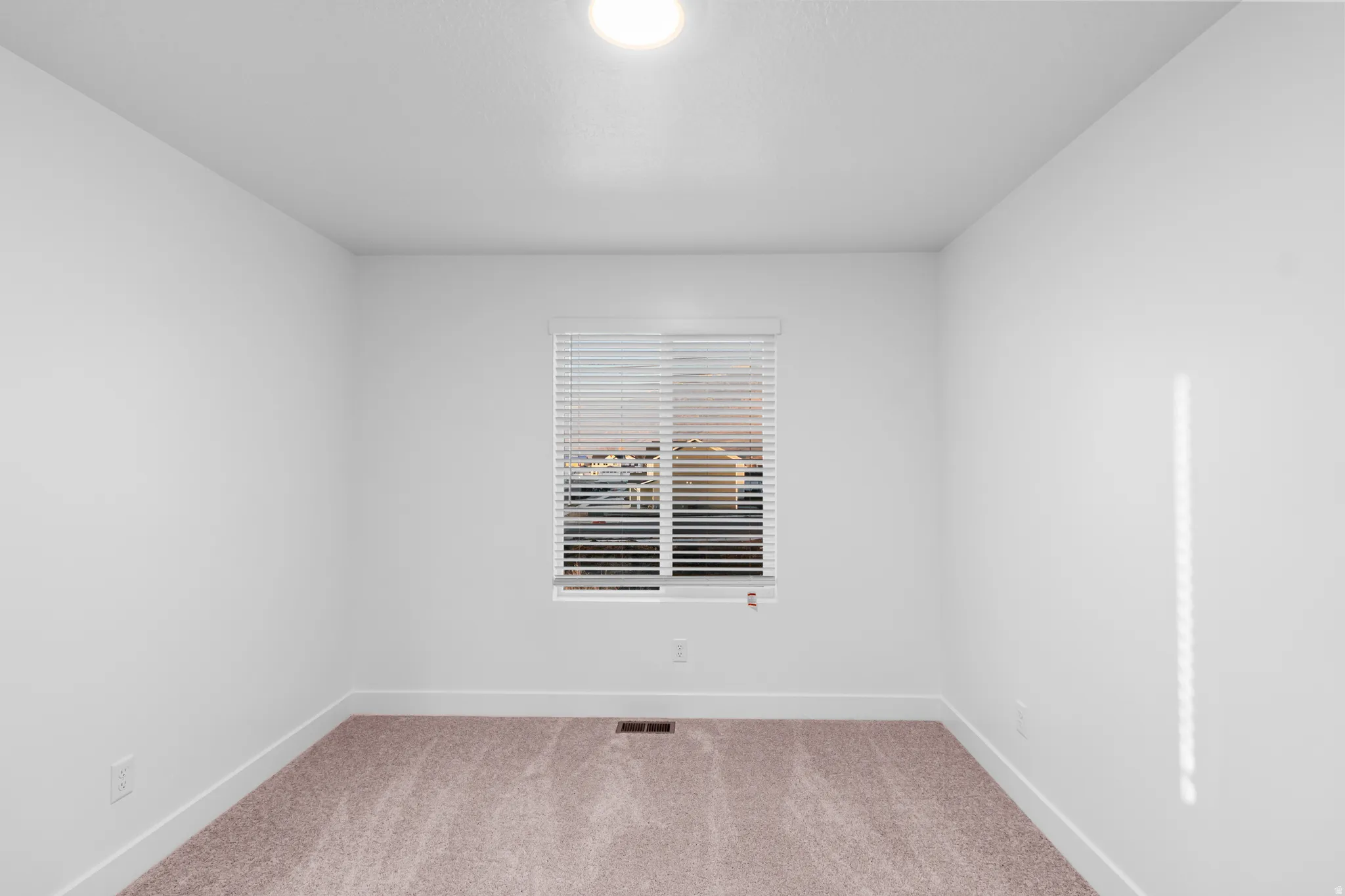 Neutral toned interior room featuring a window with horizontal blinds, overhead lighting, and carpet flooring