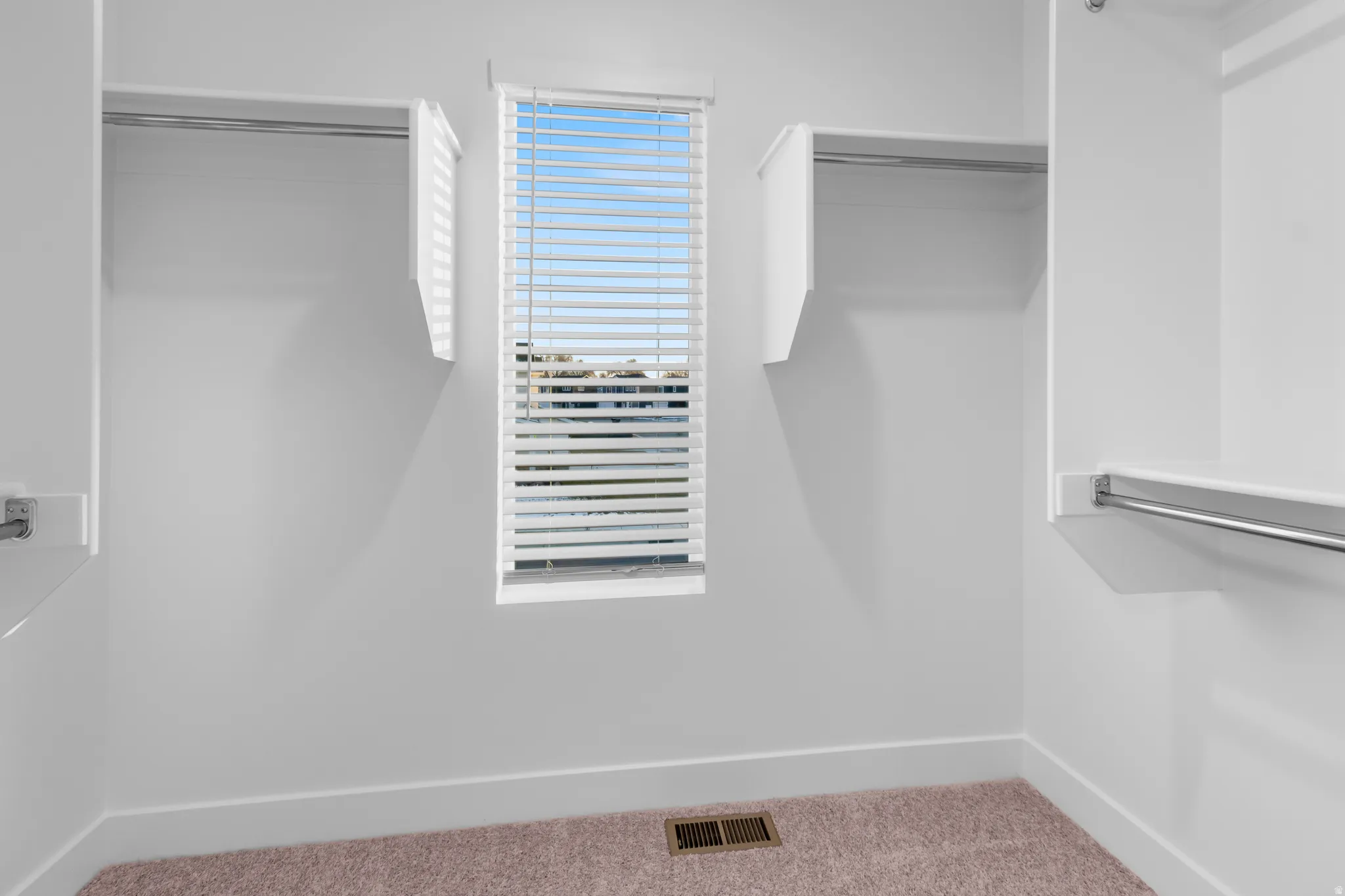 Walk-in closet featuring white built-in shelving, chrome hanging rods, a window with horizontal blinds, and neutral-toned carpeting