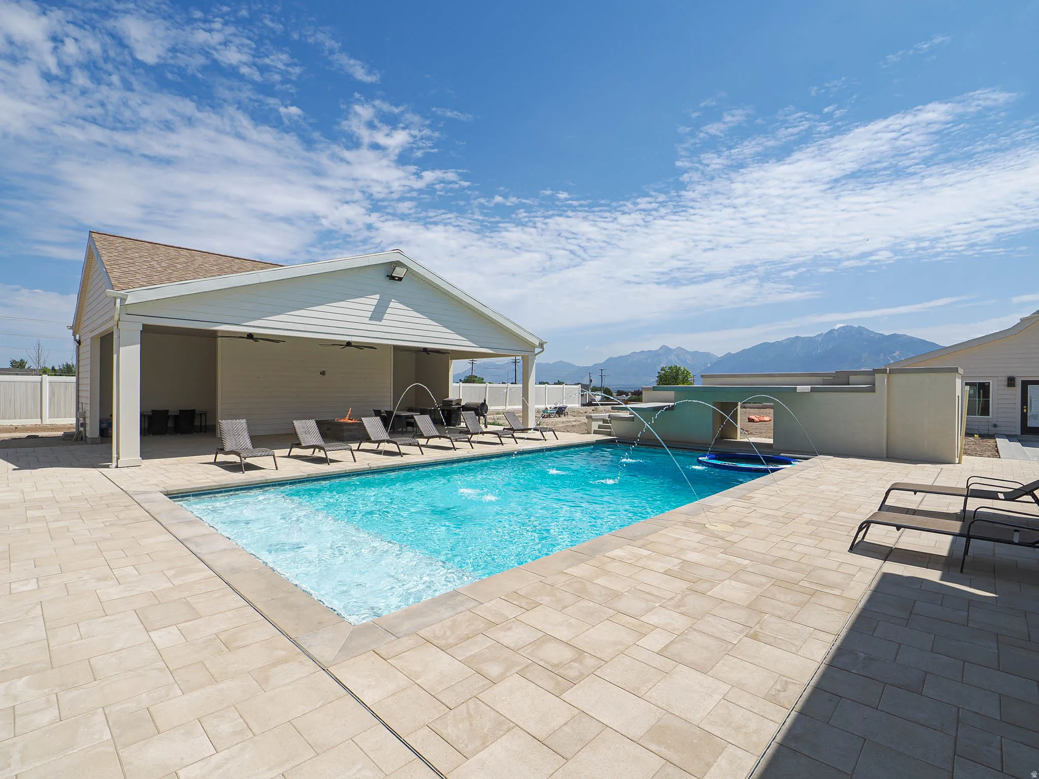 Outdoor swimming pool with integrated spa and water features, surrounded by extensive paver decking, a covered patio structure, and distant mountain scenery