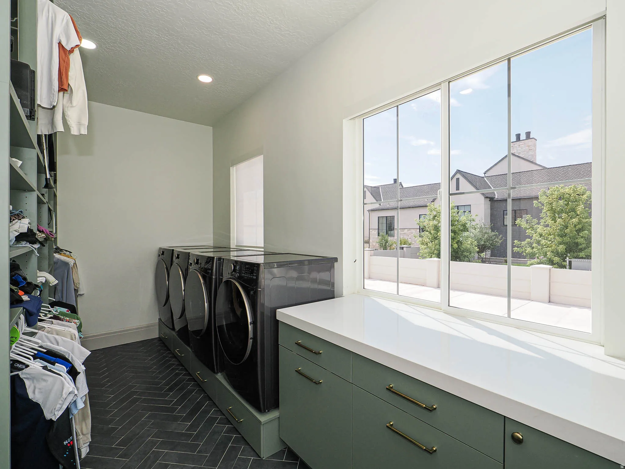 Laundry room featuring dark herringbone tile flooring, built-in shelving, two sets of front-loading appliances on integrated pedestals, and a countertop with green cabinetry