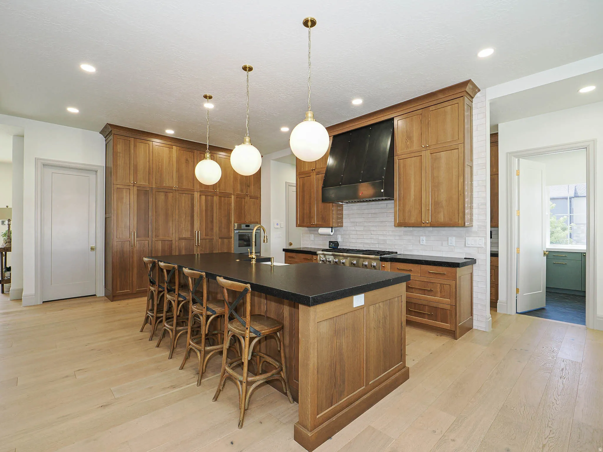 Spacious kitchen featuring a large central island with seating, extensive wood cabinetry, light wood-finish flooring, a commercial-style range with a black hood, and a light-toned brick-patterned backsplash