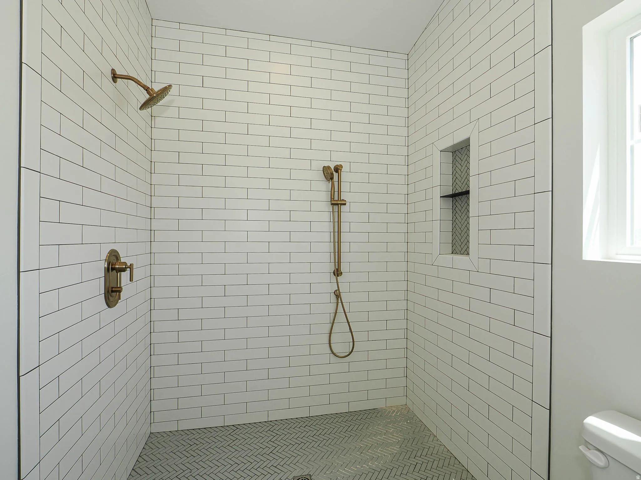 Walk-in shower featuring white subway tile walls, a herringbone pattern tile floor, and bronze fixtures including a showerhead, handheld sprayer, and control valve