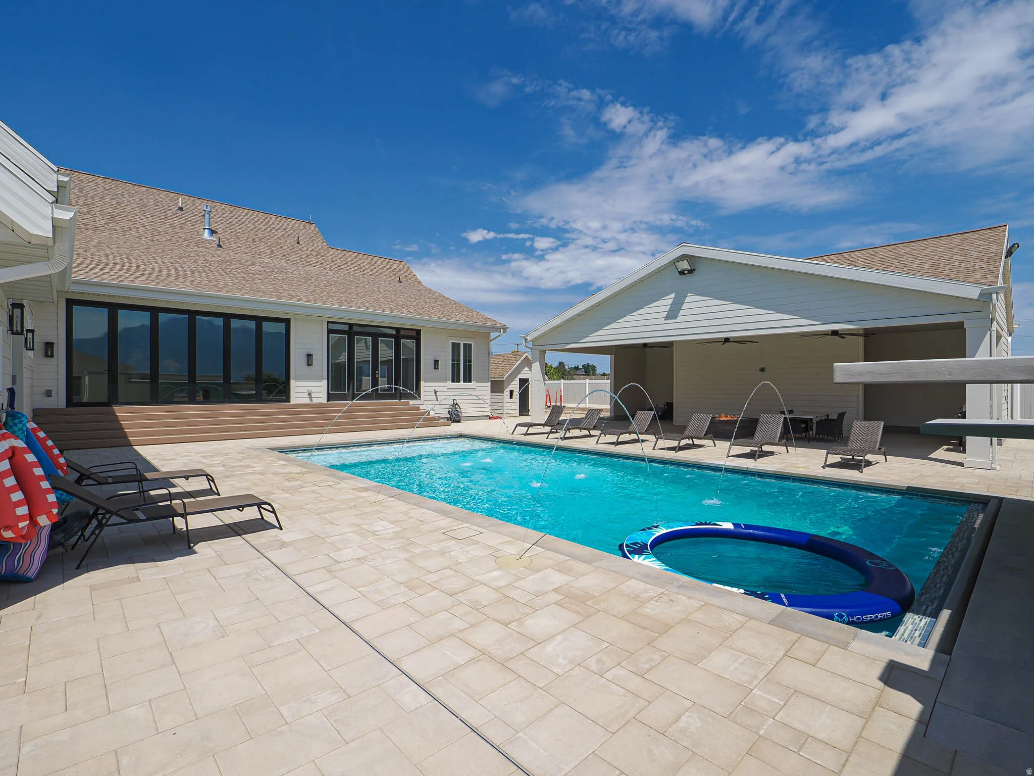 Expansive outdoor living area featuring a rectangular swimming pool with integrated water features, extensive paver patio, a covered patio structure, and a large multi-panel glass door system