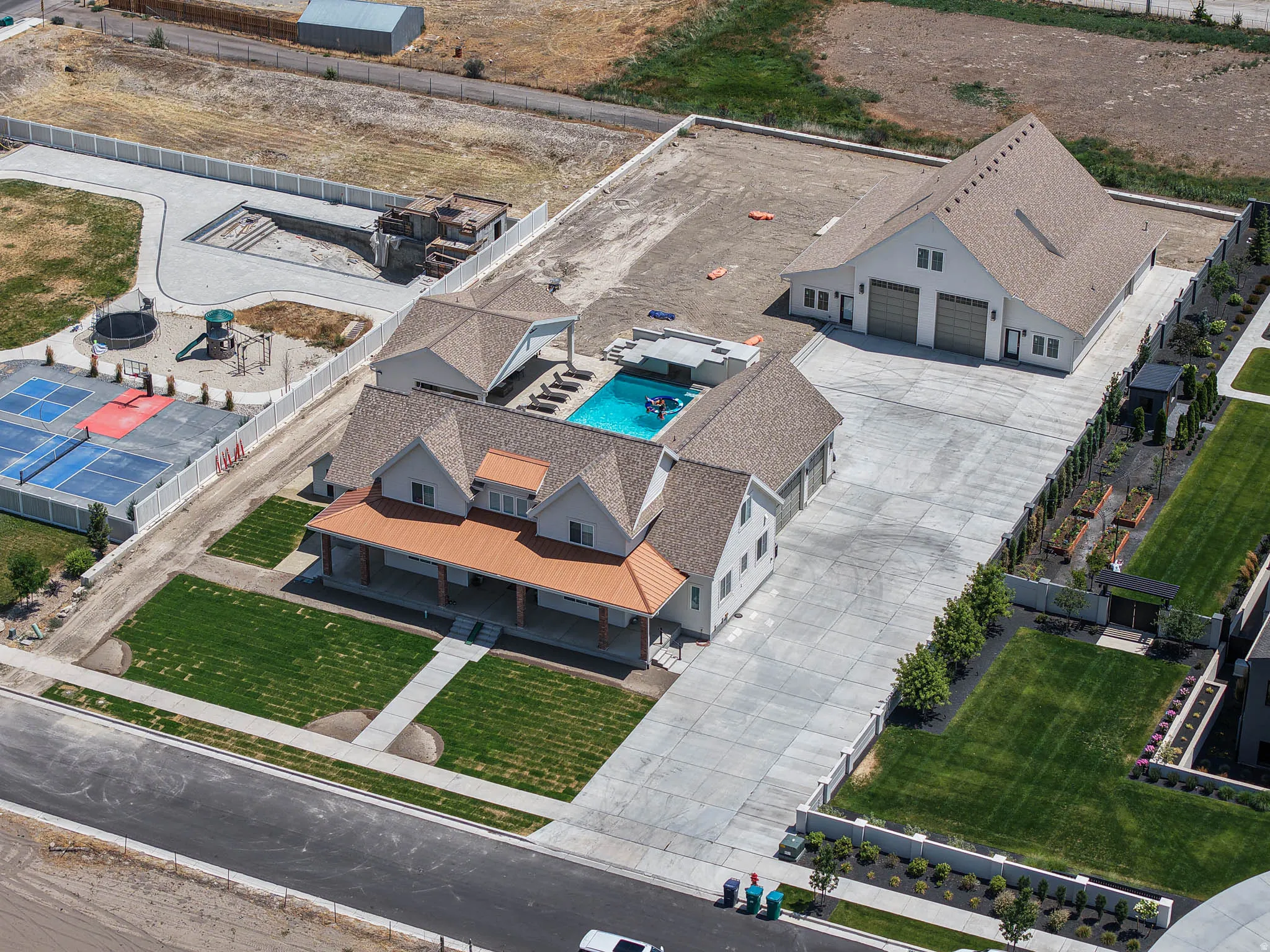 Expansive property featuring a large main residence with a covered front porch, a detached multi-car garage, a swimming pool with integrated lounge areas, a sports court, and a separate structure with an unfinished pool