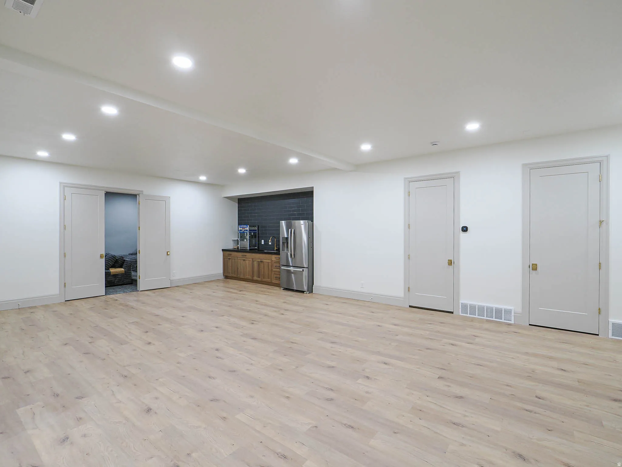 Spacious interior featuring wide plank wood-finish flooring, recessed lighting, and a built-in wet bar with a stainless steel refrigerator and dark tile backsplash