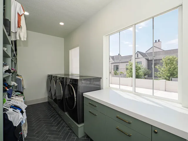 Laundry room featuring dark herringbone tile flooring, built-in shelving, two sets of front-loading appliances on integrated pedestals, and a countertop with green cabinetry