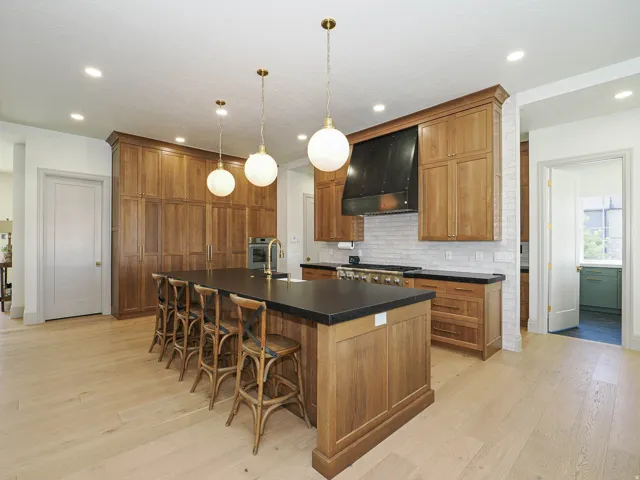 Spacious kitchen featuring a large central island with seating, extensive wood cabinetry, light wood-finish flooring, a commercial-style range with a black hood, and a light-toned brick-patterned backsplash
