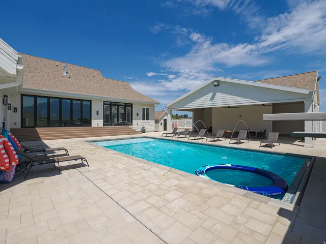 Expansive outdoor living area featuring a rectangular swimming pool with integrated water features, extensive paver patio, a covered patio structure, and a large multi-panel glass door system