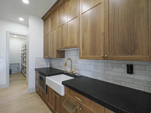 Functional utility space featuring wood-finish cabinetry, dark countertops, and a white farmhouse-style sink with a brass-finish faucet