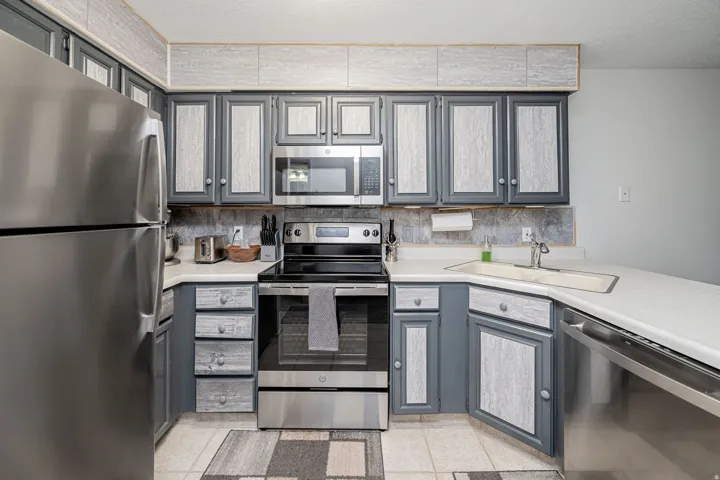 Kitchen featuring stainless steel appliances, grey and light wood-finish cabinetry, a corner sink with a stainless steel faucet, white countertops, and light tile flooring