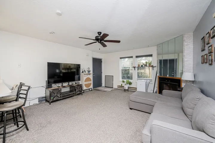 Spacious living area featuring light gray carpeting, a dark ceiling fan, multiple windows with natural light, a mirrored wall accent, and a built-in electric fireplace with a wooden mantel