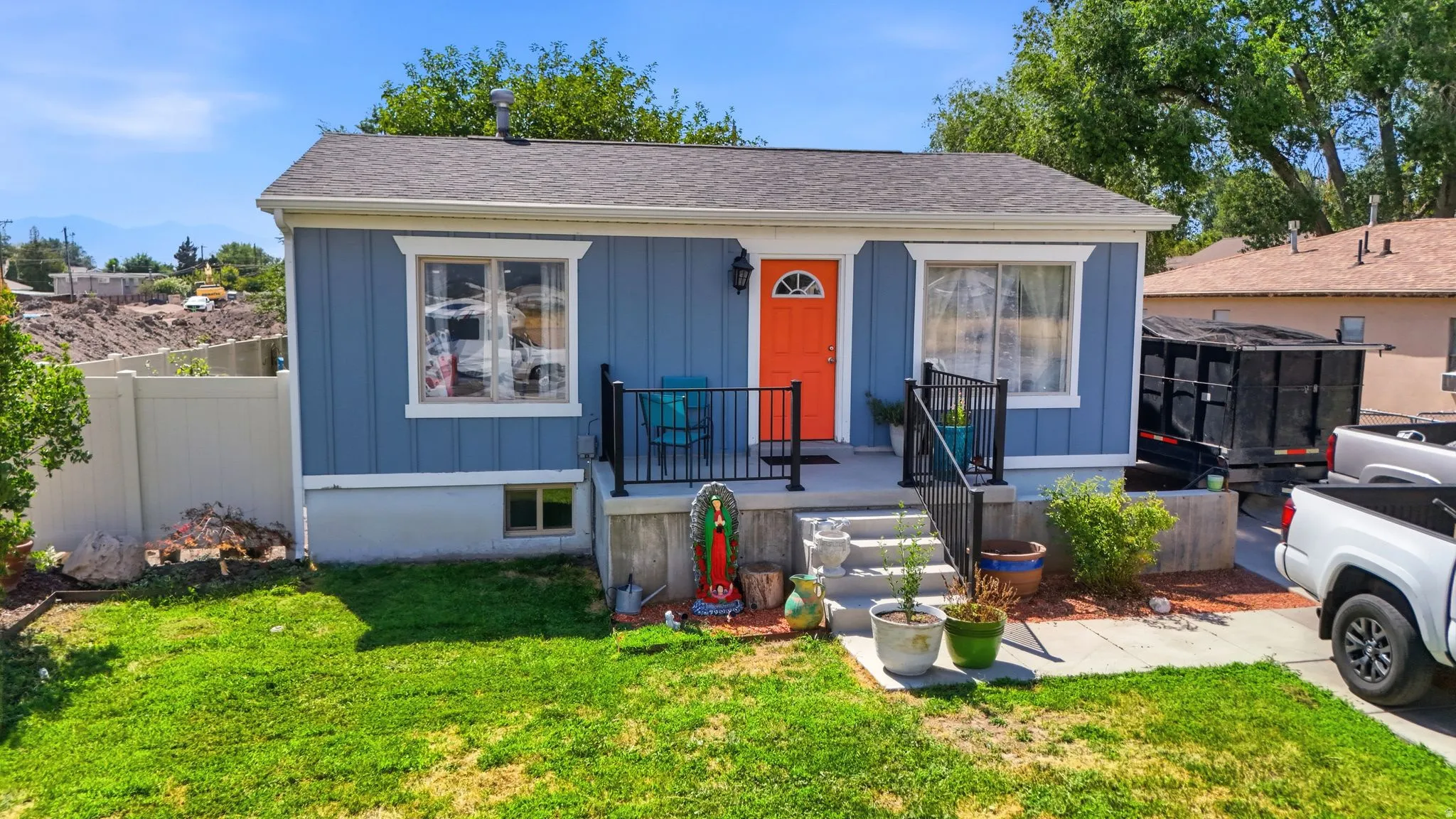 Blue exterior with white trim, featuring a front porch with black railings, a vibrant orange front door, a manicured lawn, and a white privacy fence