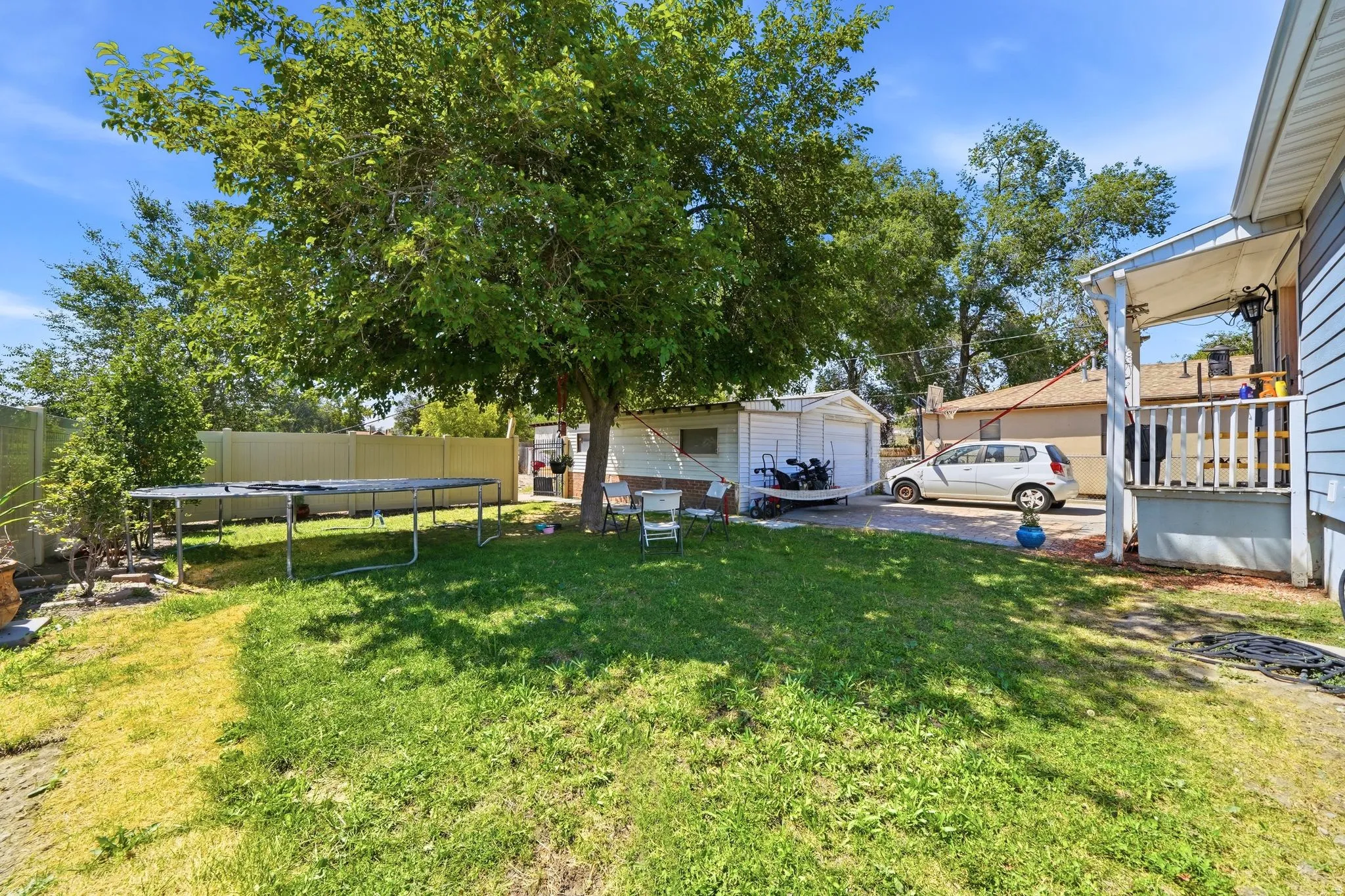 Expansive backyard featuring a mature tree providing natural shade, a white privacy fence, a concrete driveway, and a covered patio with railing