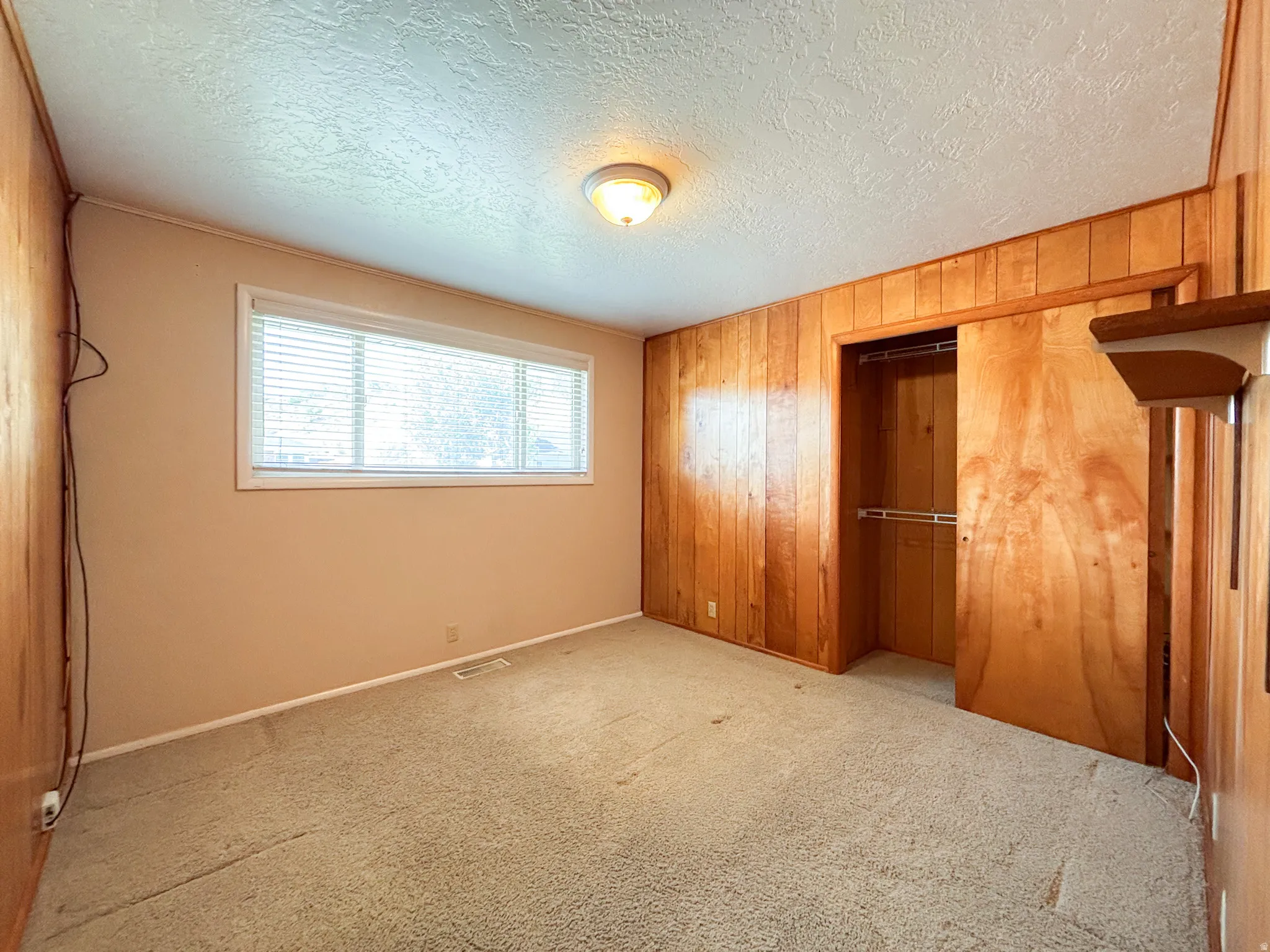 Room featuring wood paneling, carpet flooring, a single window with blinds, a built-in closet with sliding wood doors, and a ceiling-mounted light fixture