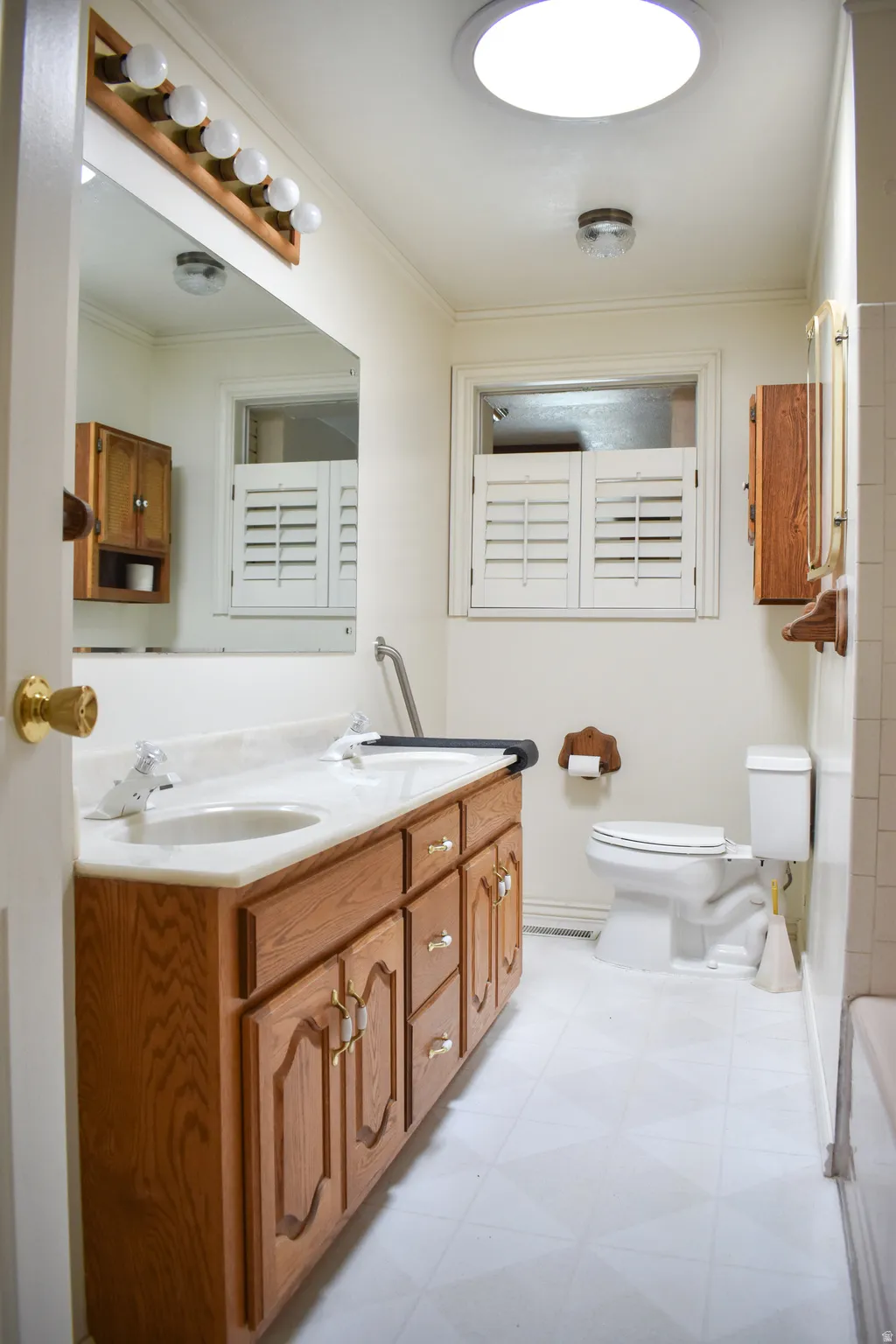 Bathroom featuring a dual-sink wood-finish vanity with a light-toned countertop, a window with white louvered shutters, and a skylight