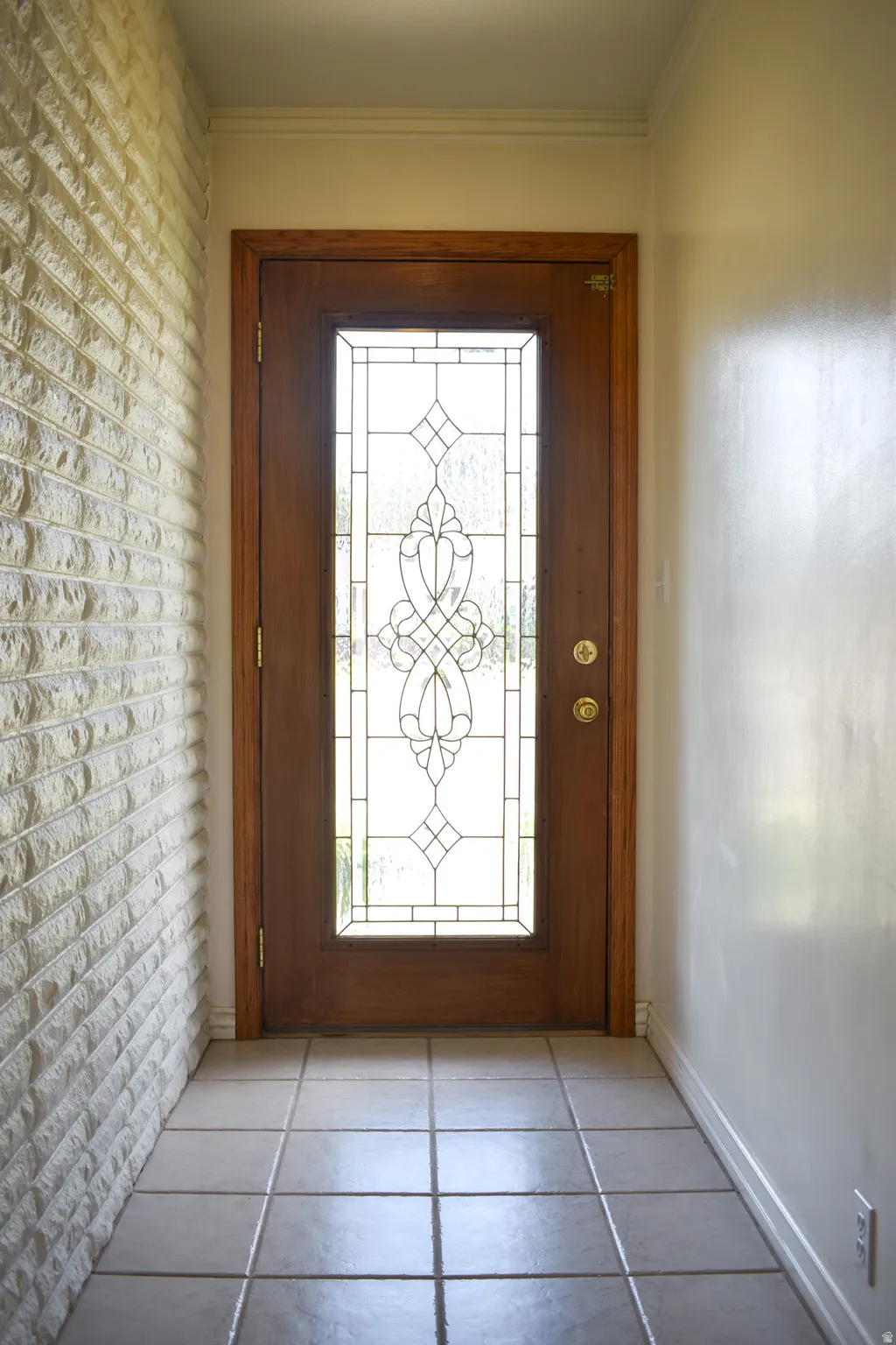 Wood-finish entry door with decorative glass insert, complemented by a textured brick-finish accent wall and square tile flooring