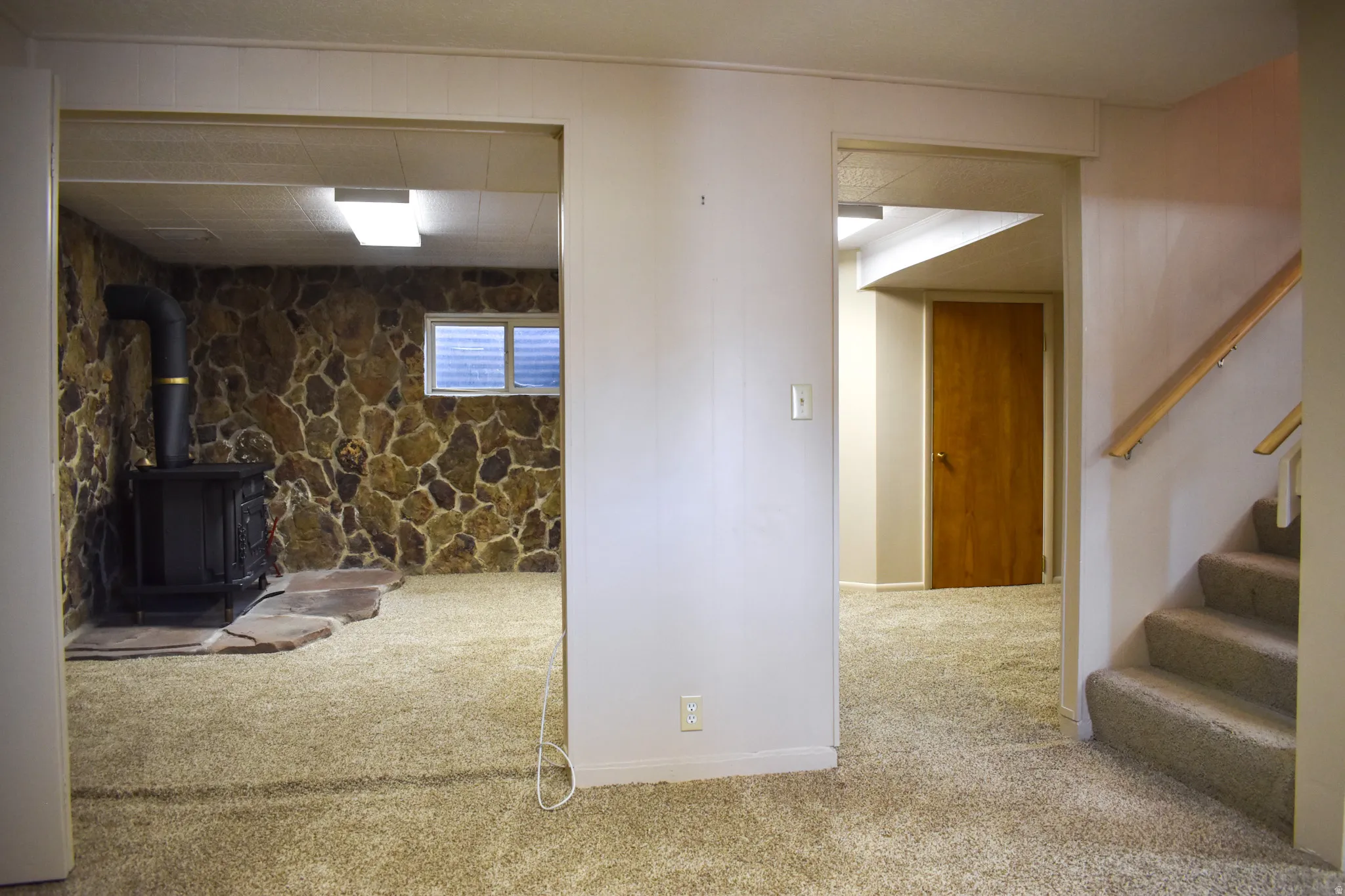 Lower level space featuring stone veneer walls, an exposed wood-burning stove on a flagstone hearth, and carpeted flooring