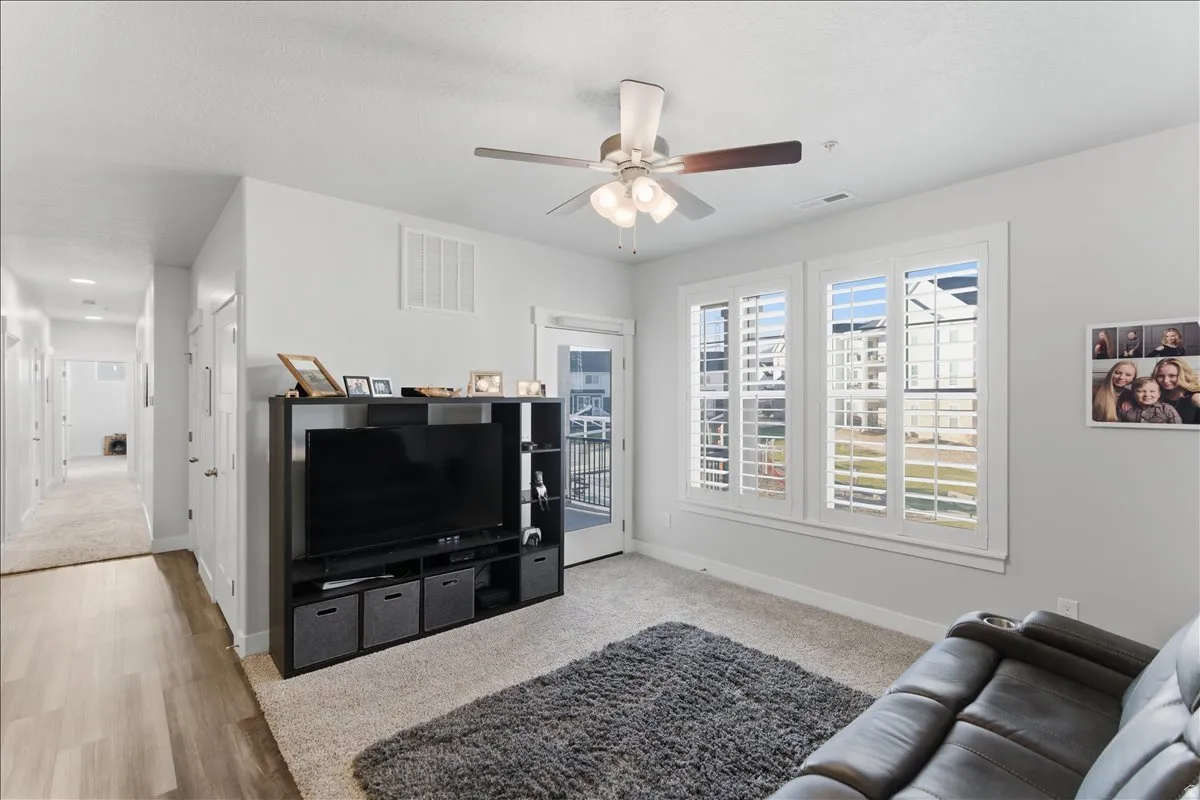 Living area featuring light gray walls, plush carpeting, white window frames with plantation shutters, a glass-paned exterior door, and a ceiling fan with integrated lighting